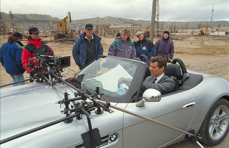 Michael Apted (ao centro), com Pierce Brosnan (ao volante) — rodagem de 