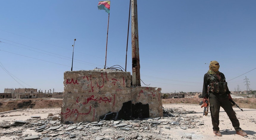 Um combatente curdo de pé com as suas armas perto de uma bandeira curda da região de Ghwairan, Síria.
